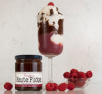 Jar of Raspberry Haute Fudge on the left of a raspberry hot fudge sundae with a marble wall behind it all. On the right of the sundae is a small glass bowl of raspberries and a few loose raspberries on the countertop