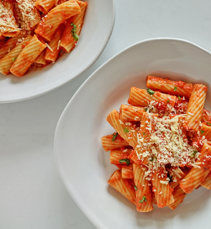 Two white ceramic plates of tortiglioni pasta covered in vodka sauce, bits of herbs, and shredded parmesan cheese
