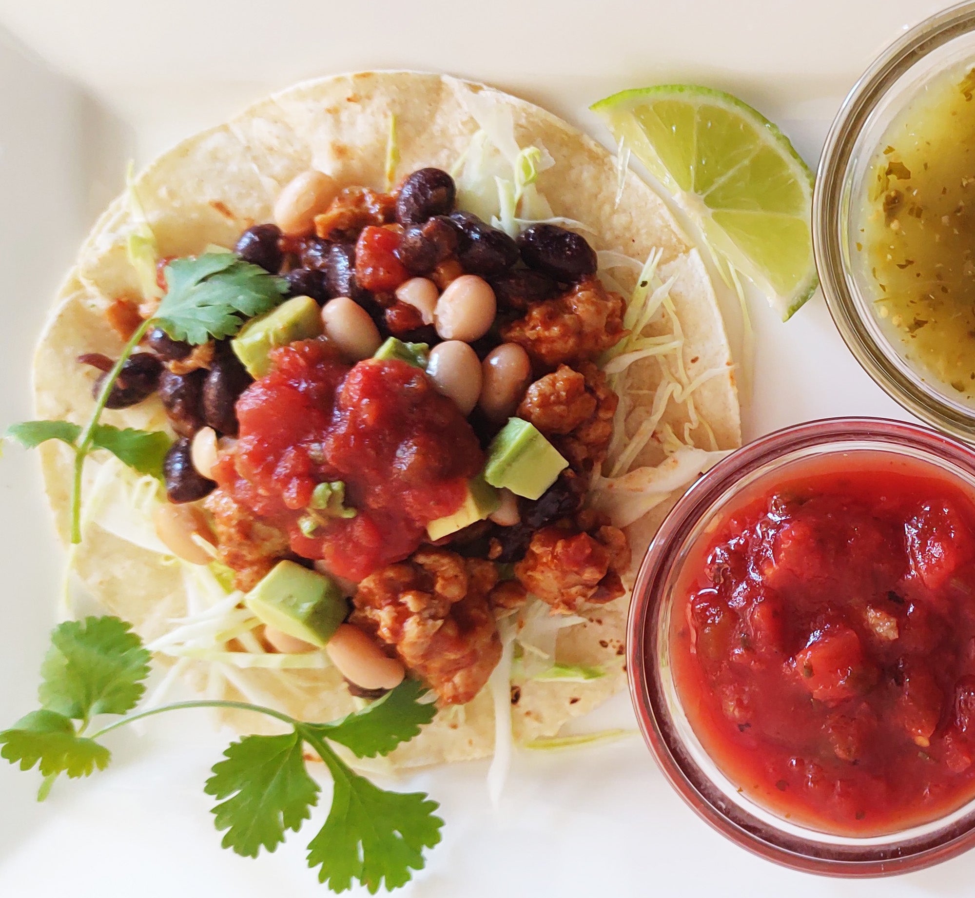 Overhead view of an open-face taco on square white plate garnished with cilantro. Next to the taco is a slice of lime, a glass bowl of salsa verde, and a glass bowl of salsa roja.
