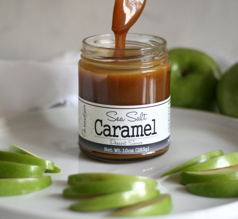 Unlidded jar of sea salt caramel sauce on a white plate surrounded by thin slices of green apple, with two full apples sitting behind the plate. A spoon is above the unlidded jar, drizzling caramel back down into it