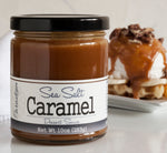 Jar of sea salt caramel sauce in the foreground. In the background, there is a waffle topped with scoop of vanilla ice cream, topped with sea salt caramel and cookie bits, all on a white plate.