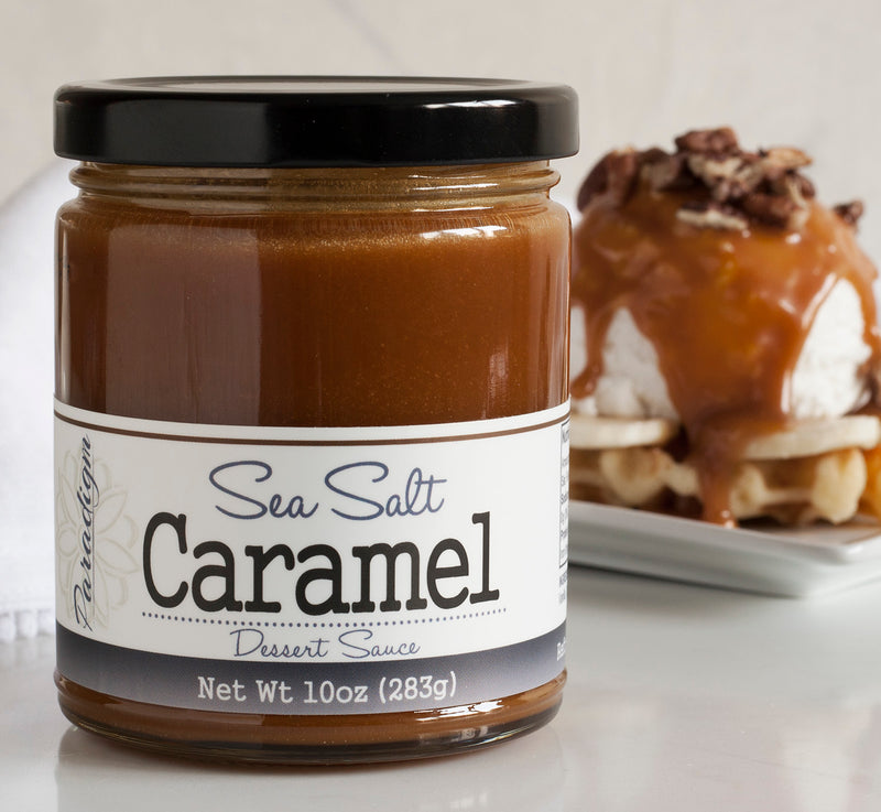 Jar of sea salt caramel sauce in the foreground. In the background, there is a waffle topped with scoop of vanilla ice cream, topped with sea salt caramel and cookie bits, all on a white plate.
