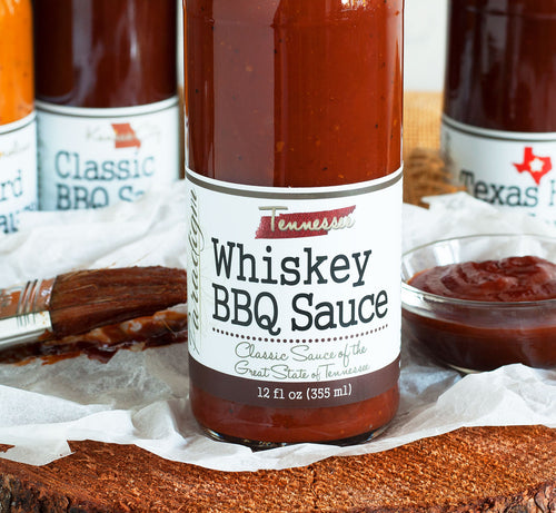 Bottom half of bottle full of whiskey BBQ sauce on crinkled wax paper on top of wood round. Behind the bottle is a glass bowl of BBQ sauce, a couple more bottles of BBQ sauce, and a basting brush covered in BBQ sauce.