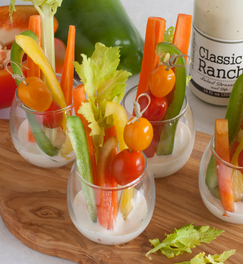 Wooden cutting board on top of which are four glass cups filled with slices of bell pepper, celery stalks, and skewers of cherry tomatoes. Behind the cutting board is a bottle of Classic Ranch, a green bell pepper, and a red bell pepper. 