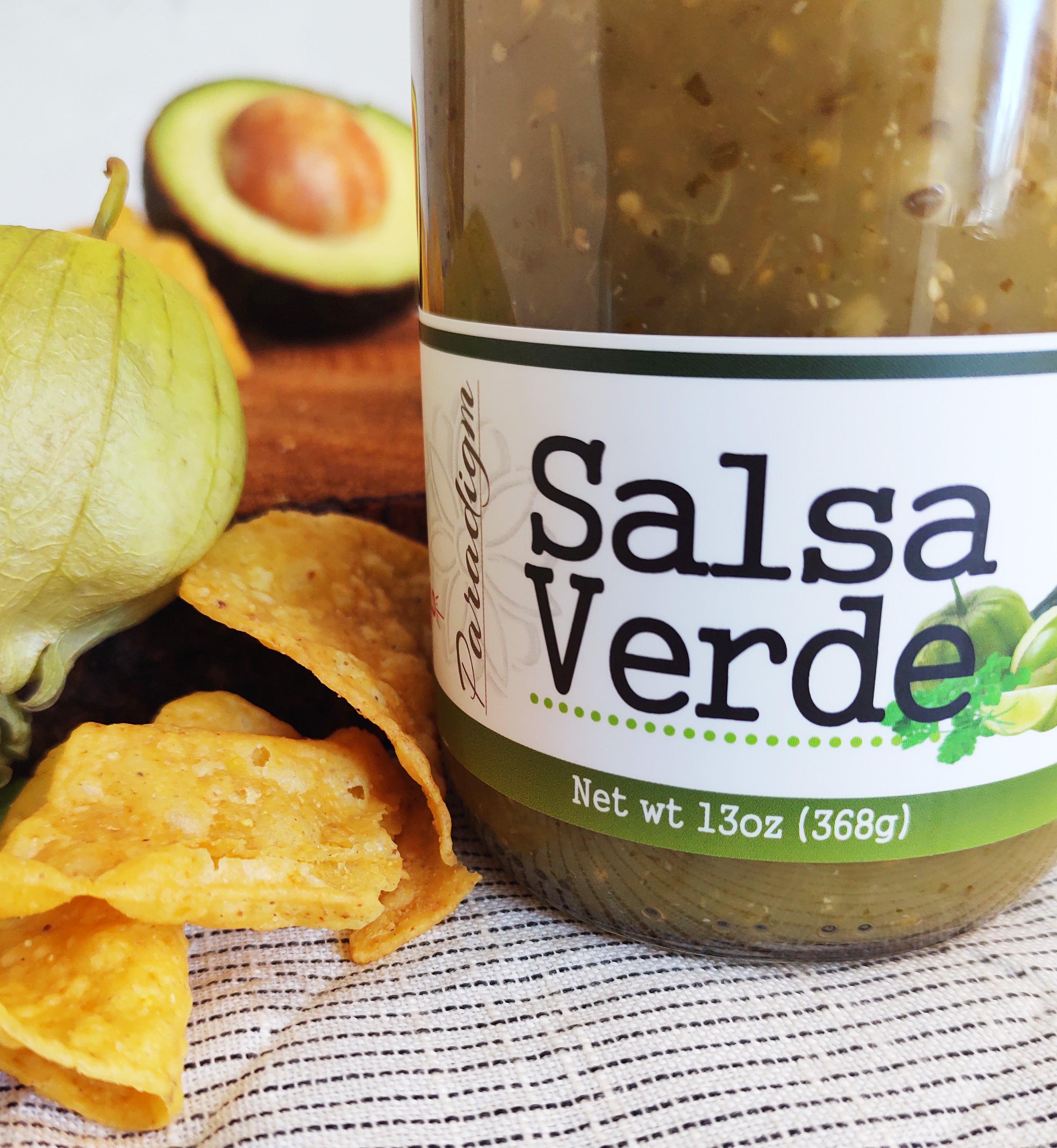 Closeup of jar of salsa verde on black and white striped linen napkin. Next to the jar are a few tortilla chips, a pepper, and half an avocado.