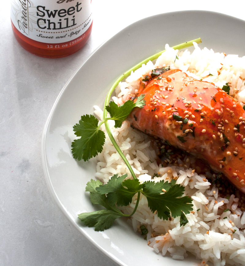 Slab of salmon covered in thai sweet chili sauce and sesame seeds on top of a bed of rice and garnished with a piece of cilantro sitting on a white ceramic plate next to a bottle of Thai Sweet Chili.