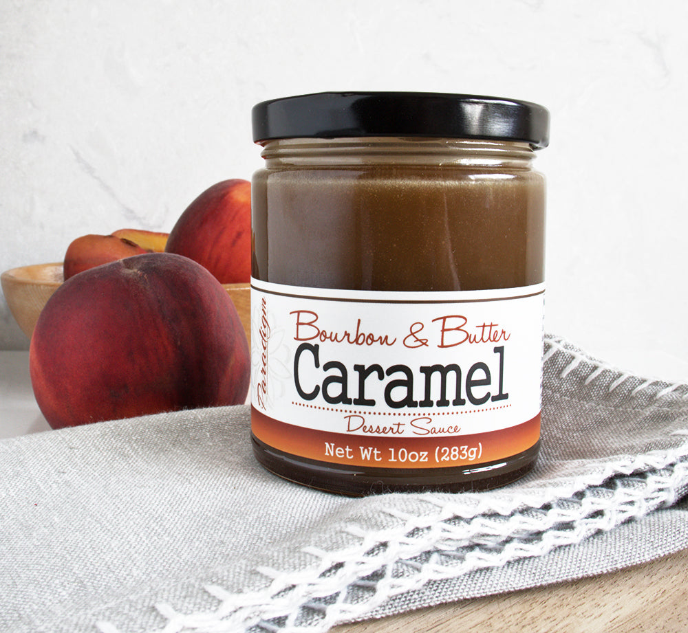 Jar of Bourbon & Butter Caramel Dessert Sauce sitting on top of a grey linen napkin with peaches behind the jar