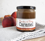 Jar of Bourbon & Butter Caramel Dessert Sauce sitting on top of a grey linen napkin with peaches behind the jar