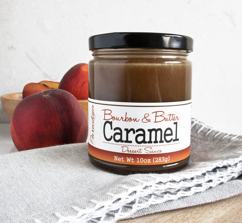 Jar of Bourbon & Butter Caramel Dessert Sauce sitting on top of a grey linen napkin with peaches behind the jar