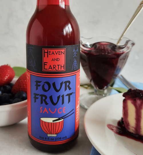 Closeup of bottle of Four Fruit Sauce in front of decorative glass filled with four fruit sauce, white bowl blueberries and strawberries, and a piece of berry cheesecake.