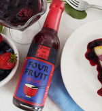 Bottle of Four Fruit Sauce laying on countertop next to plate of berry cheesecake. By the top of the bottle is a fork and leaves, and to the side is a small bowl of strawberries and blueberries and a decorative glass of fruit sauce with whole berries in it.