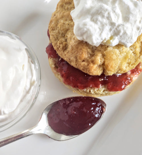 Biscuit with jam topped with whipped cream on a white plate. Next to the biscuit on the plate is a small glass bowl of whipped cream and a spoon full of jam.