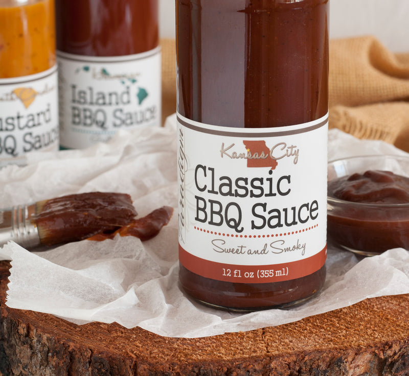 Bottom half of bottle full of Classic BBQ sauce on crinkled wax paper on top of wood round. Behind the bottle is a glass bowl of BBQ sauce, a couple more bottles of barbecue sauce, and a basting brush covered in BBQ sauce.