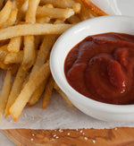 Closeup of french fries and ceramic white bowl of ketchup on top of tissue paper, spread over a wooden cutting board.