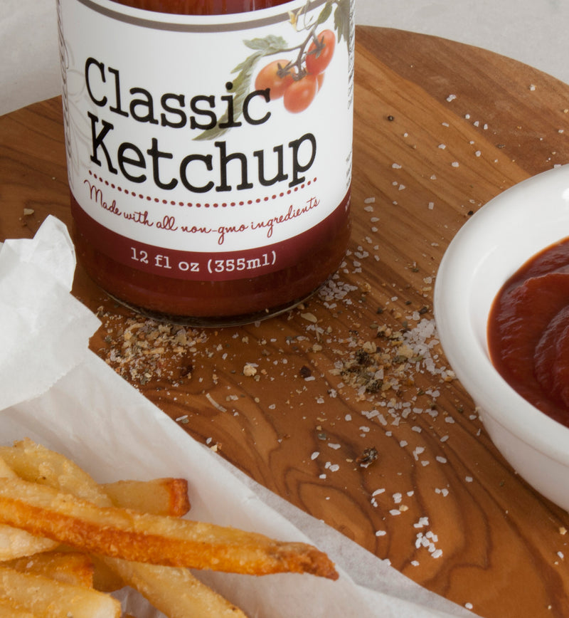 Bottom half of bottle of Classic Ketchup on wood cutting board sprinkled with spices. A small ceramic white bowl of ketchup and a piece of tissue paper covered in French fries sit on the board near the bottle.