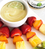 Small ceramic white bowl full of lime curd with half circle slice of lime on a white plate of fruit skewers containing a strawberries, peaches, pineapple, and kiwi. Next to the plate is a jar of lime curd