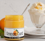Unlidded jar of Orange Curd with a spoon in it in front of an orange and two leaves. To the right of the jar is a glass sundae cup filled with vanilla ice cream and orange curd on a white plate on top of a folded white linen napkin