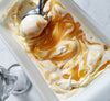 Rectangular dish filled with vanilla ice cream with orange curd mixed into it and an ice cream scoop scooping out a portion of the ice cream. Next to the dish is an empty glass sundae cup