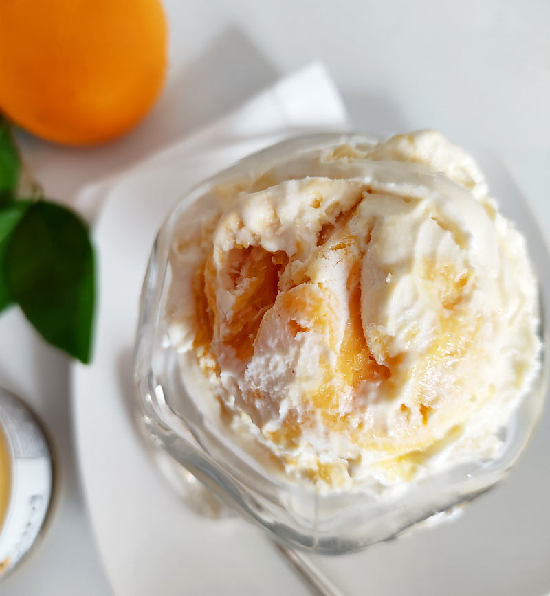 Overhead view of scoop of vanilla ice cream with orange curd mixed into it, held in a glass sundae cup. Under the sundae is a white plate on top of a white linen napkin. Next to the napkin are an orange, a few leaves, and a jar of Paradigm Orange Curd