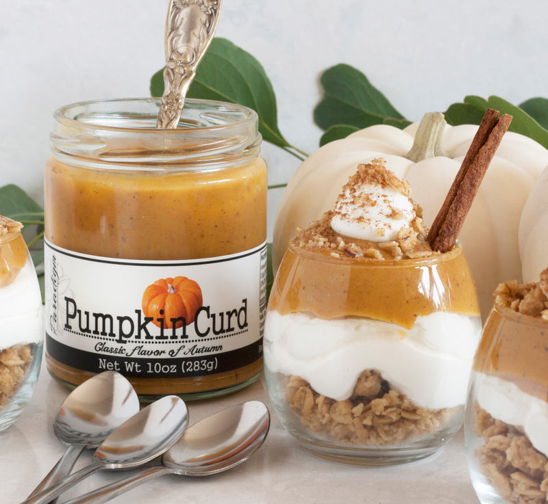 Unlidded jar of Pumpkin Curd with spoon in it in line flanked by three pumpkin curd parfaits, each topped with cool whip, brown sugar, and sticks of cinnamon. Three spoons lay in the foreground and white pumpkins and a few leaves sit behind the parfaits. 