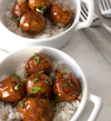 Two white ceramic bowls with handles filled with white rice and meatballs covered in Polynesian Teriyaki sauce and green onions. In the top right corner are two forks on a gray linen napkin.