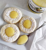 Thumbprint cookies topped with powdered sugar and lemon curd on a white square plate with a spoon full of lemon curd. The plate sits on a piece of crinkled tissue paper next to a jar of Paradigm Lemon Curd